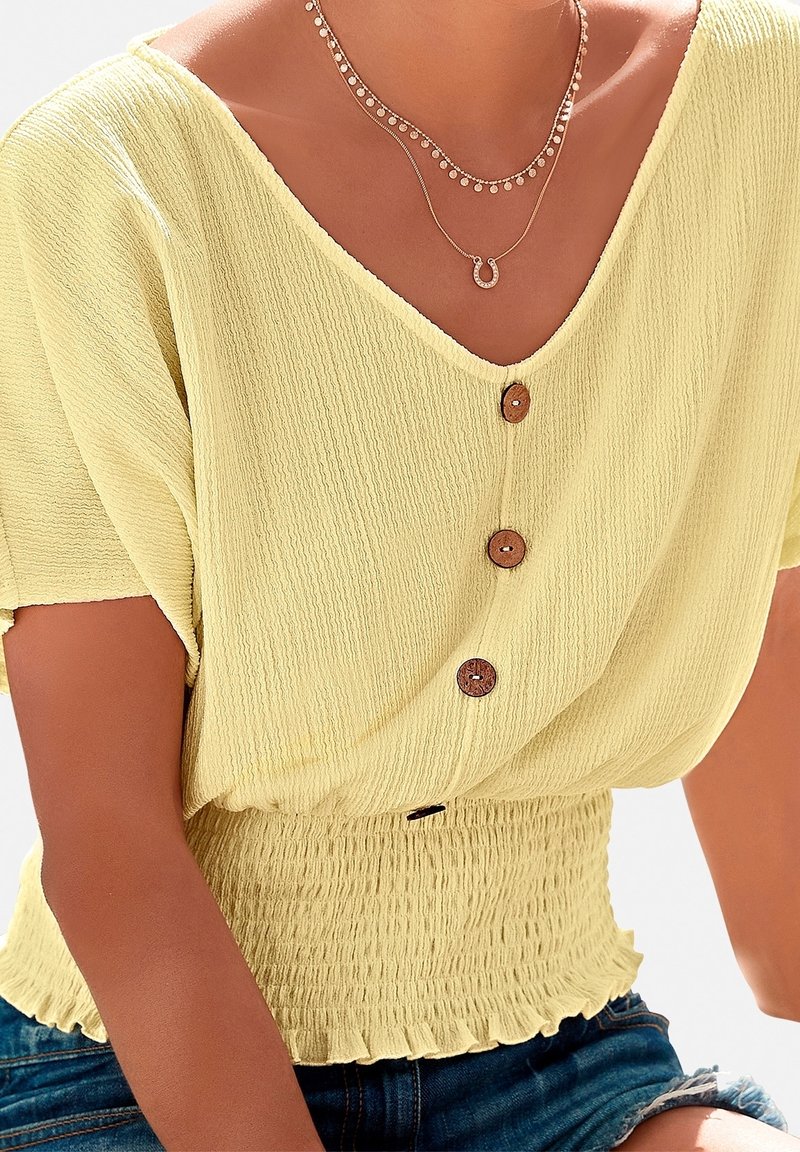 Femme portant un haut texturé jaune clair boutonné avec une taille smockée et des colliers dorés superposés, assorti à un short en jean déchiré.