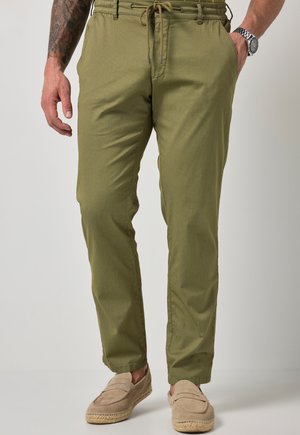 Mand iført olivengrønne bukser med snøre og beige slip-on sko, stående med den ene hånd i lommen og synlig tatovering på venstre arm.