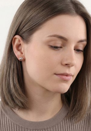 Jeune femme aux cheveux bruns et raides portant des boucles d'oreilles clous rondes en argent et un pull côtelé taupe, regardant vers le bas avec une expression neutre.