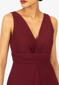 Bordeauxfarbenes Kleid mit tiefem V-Ausschnitt, gefälteltem Oberteil und geraffter Taille. Glatter Stoff mit einem fließenden Rockdetail für elegante Bewegungen.