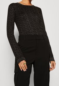 Czarny longsleeve z teksturowanym wzorem i metalicznymi detalami, zestawiony z czarnymi spodniami z wysokim stanem. Na dłoni srebrny pierścionek.