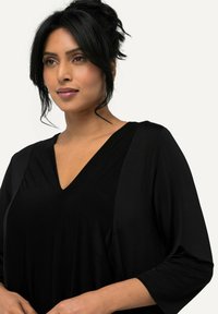 Top nero a scollo a V con maniche svasate 3/4, realizzato in un tessuto morbido, caratterizzato da dettagli drappeggiati e una vestibilità rilassata. Texture liscia e leggera lucentezza.