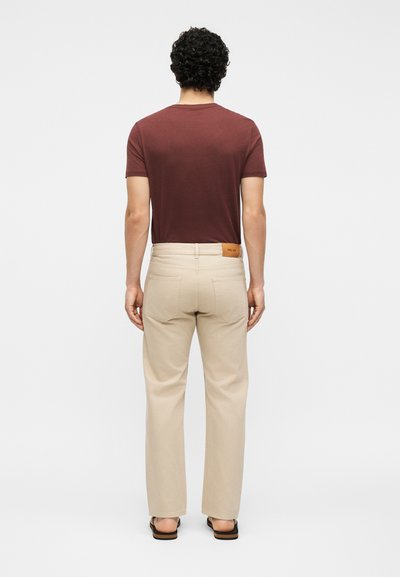 Homme aux cheveux bouclés et foncés portant une chemise marron à manches courtes, un pantalon beige avec des poches à l'arrière et des sandales noires, debout de dos.