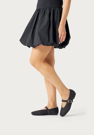 Donna che indossa una gonna corta nera a bolle e ballerine nere intrecciate Mary Jane con fibbia a doppio anello, in piedi con le gambe incrociate su uno sfondo chiaro.