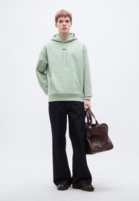 Φωτεινό πράσινο hoodie με τσέπη στο μπροστινό μέρος και μαύρο λογότυπο, συνδυασμένο με σκούρο μπλε τζιν φαρδιάς γραμμής και καφέ δερμάτινη τσάντα ώμου.