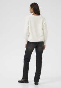 Hvid strikket sweater med en afslappet pasform, båret med mørke denim jeans og sorte loafers. Bagfra ses ribbestrikket kant og sømdetaljer.