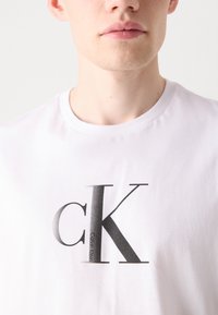 Balta medvilninė marškinė su apvaliu kaklu, ant krūtinės puikuojasi didelis juodas "cK" logotipas šiuolaikišku šriftu. Minkšta tekstūra.