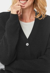 Schwarzer gestrickter Cardigan mit V-Ausschnitt. Verfügt über eine gerippte Textur und einen auffälligen weißen Knopf an der Vorderseite. Lange Ärmel sichtbar.