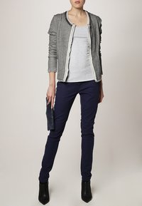 Chaqueta gris texturizada con cremallera al frente y cuello adornado, combinada con una camiseta gris ajustada, pantalones azul marino y botines negros de tacón.