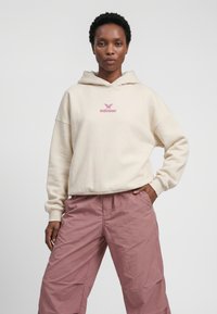 Vrouw draagt een crèmekleurige hoodie met een roze vlinderlogo en tekst, en losse roze broek, staand met één hand in de zak tegen een effen achtergrond.