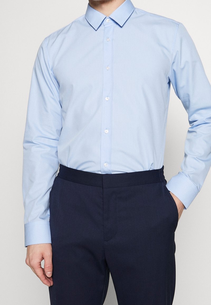 Homme portant une chemise bleue claire boutonnée rentrée dans un pantalon bleu marine foncé, une main dans la poche, sur un fond clair uni.