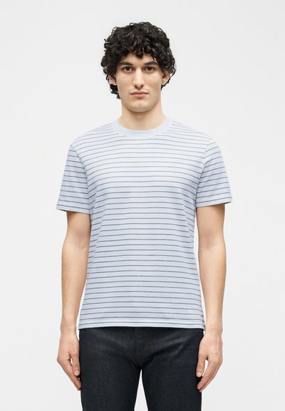 Άνδρας με σγουρά μαλλιά που φοράει ένα ανοιχτό μπλε κοντομάνικο μπλουζάκι με λεπτές οριζόντιες ρίγες και σκούρο παντελόνι, που στέκεται μπροστά σε απλό φόντο.