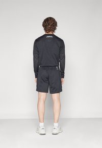 Crna dugaa majica za sport s mrežastom teksturom i logom, u kombinaciji s crnim kratkim hlačama. Bijele tenisice upotpunjuju odjevnu kombinaciju.