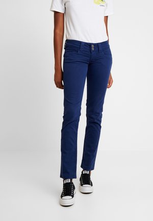 Persoon draagt marineblauwe aansluitende broek met dubbele knoopsluiting, wit grafisch T-shirt en zwarte hoge sneakers, staand op een effen achtergrond.