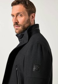 Uomo con barba che indossa una giacca nera con colletto alto e tasca centrale con zip, rivolto a sinistra contro uno sfondo chiaro e semplice.