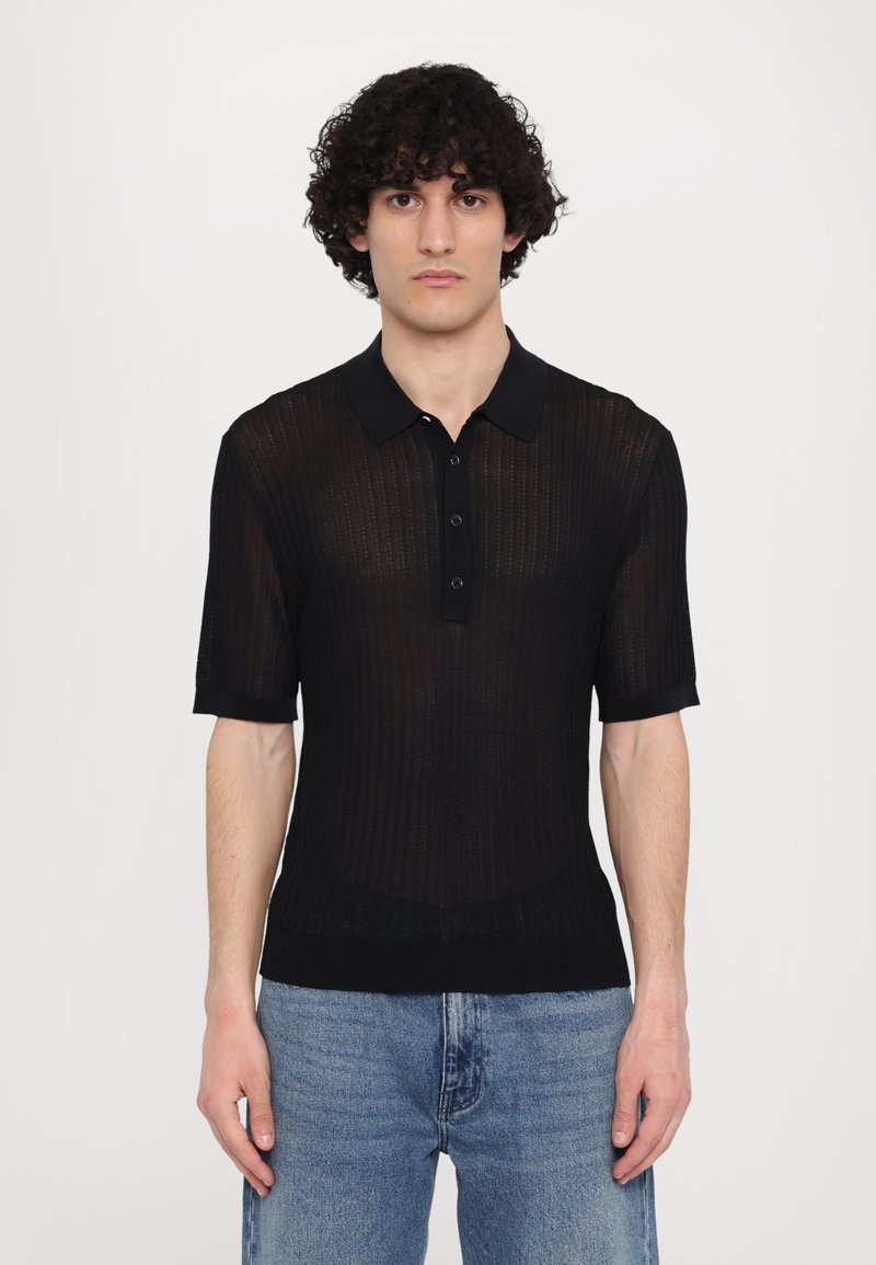 Fekete, rövid ujjú pólóing, bordás textúrával, három gombos kivágással és gallérral. Kék farmernadrággal párosítva.