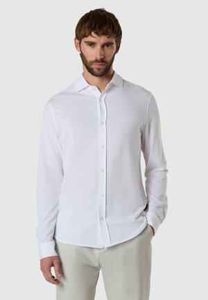 Uomo con capelli castani corti e barba, che indossa una camicia bianca a maniche lunghe con bottoni e pantaloni beige chiaro, in piedi con una mano in tasca.