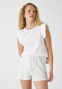 Witte top met korte mouwen en ruche schouderdetails, gecombineerd met witte textuurshorts met een koord. Beide zijn gemaakt van zachte, lichte stoffen.