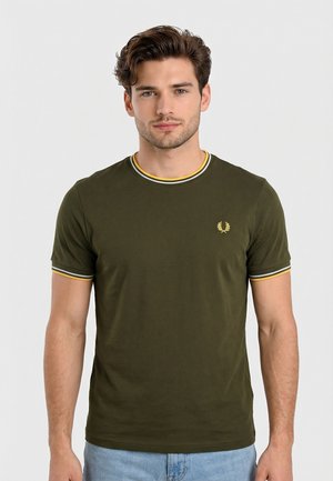 Jonge man met kort krullend haar, gekleed in een olijfgroen T-shirt met gele en witte biezen aan mouwen en kraag, staande voor een effen achtergrond.