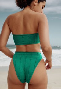 Zelený bandeau vršek bikin s vertikálním žebrováním a odpovídajícími vysokými kalhotkami; texturovaná látka; minimalistický design; pohled zezadu.