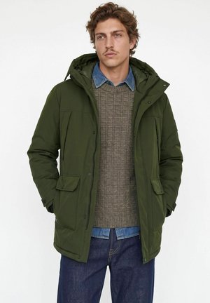 Hombre con chaqueta de invierno con capucha de color verde oliva sobre un suéter gris de punto y camisa vaquera azul, con vaqueros azules, de pie frente a un fondo blanco.