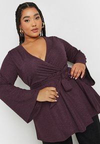 Top envolvente morado con mangas largas de campana, tejido texturizado y detalle de cintura con lazo, con un diseño de escote en V profundo.