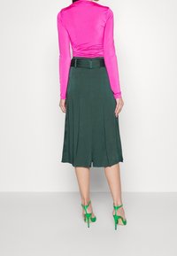 En figurnära långärmad rosa topp i kombination med en svängande grön A-linjeformad kjol och gröna högklackade sandaler, som visar en slät struktur och plisserad design.