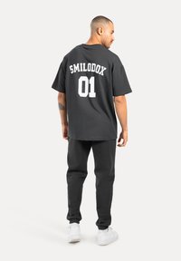 Czarna, oversize t-shirt z białym nadrukiem "SMILODOX 01" na plecach, w połączeniu z ciemnymi dresami i białymi sneakersami.