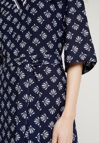 Marinblå klänning med ett vitt fläktmönster, korta ärmar och en knytbar midja för en figursydd look. Slät tygtextur.