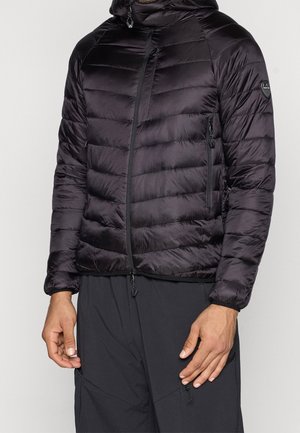 Homme portant une doudoune noire matelassée avec capuche et poches zippées, associée à un pantalon noir sur fond uni.