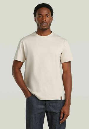 Man with short curly hair wearing a plain beige short-sleeve t-shirt and dark blue jeans standing with one hand in pocket.