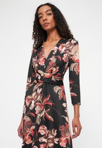 Langsleeve jurk van zwarte stof met een bloemenpatroon in rood, beige en bruin, met een V-hals en een wikkeltaille ontwerp.