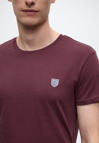 Sügavpunane ümmarguse kaelusega t-särk, valmistatud siledast puuvillast. Vasakul rinnal on väike valge kilbi kujuline logo. Lühikesed varrukad.