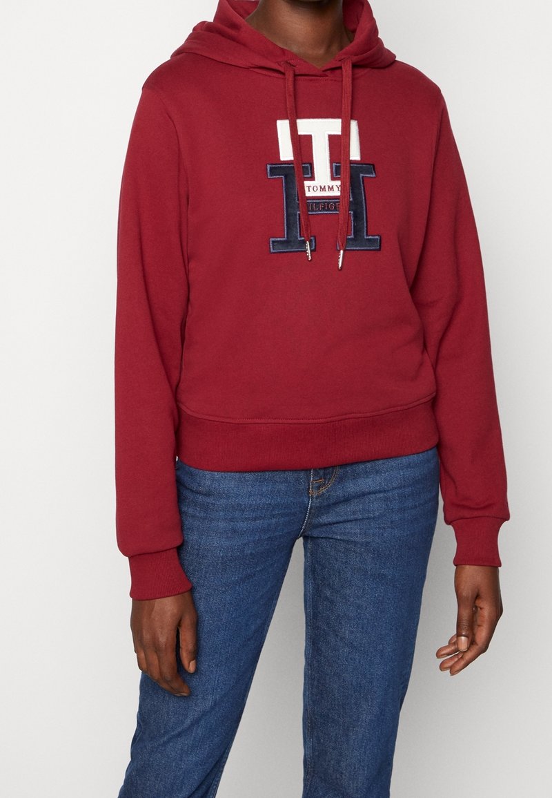 Sudadera color burdeos con un logo en tonos azul marino y blanco, cordones en el cuello y puños y dobladillo acanalados. Se combina con unos vaqueros azules.