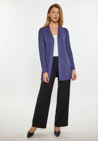 Cárdigan largo morado hecho de una tela suave, diseño de frente abierto, usado sobre una camisa blanca y combinado con pantalones de pierna ancha negros y tacones.
