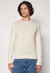 Light beige knit sweater with long sleeves and a crew neckline. Features ribbed cuffs and hem, paired with blue denim jeans.