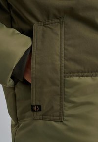 Olivgrön vadderad jacka med en bröstficka som har en vikbar klaff. Texturerad tyg och sömnadsdetaljer nära fickan. Varumärkesetikett synlig.