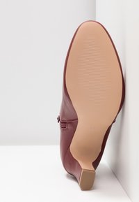 Chaussure à talon haut en cuir bordeaux, posée à l'envers dans un coin blanc, affichant une semelle beige texturée et un talon carré.