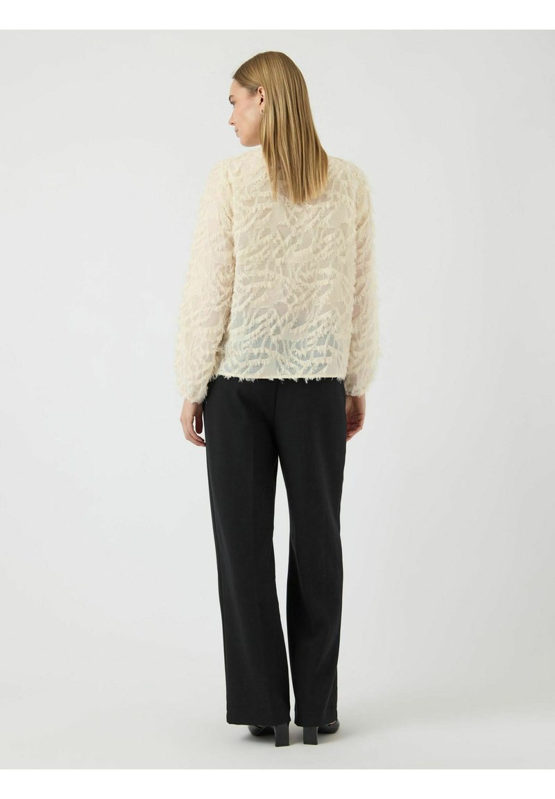 Cremfarbener, strukturierter Oberteil mit langen Ärmeln und gewelltem Muster, kombiniert mit schwarzen, geraden Hosen. Einfaches Design, minimale Hardware.