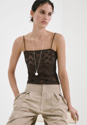 Femeie purtând un top negru din dantelă cu bretele subțiri, pantaloni bej plisati și un colier lung cu un pandantiv rotund, stând așezată cu mâinile în buzunare.