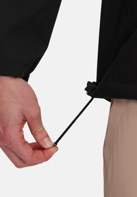 Zwarte jas met een hand die een verstelbaar elastisch koord aan de onderkant trekt. Effe textuur, minimalistisch ontwerp en een getailleerde silhoutte.