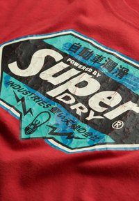 Camiseta roja de algodón con un parche de logo desgastado en azul y negro con texto blanco, rayos y un gráfico de bombilla.