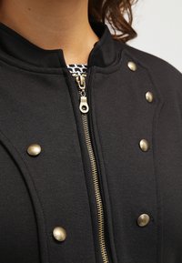 Czarny sweter z zamkiem błyskawicznym, z wysokim kołnierzem, mosiężnymi zatrzaskami oraz teksturowanym materiałem z widocznymi detalami szycia.