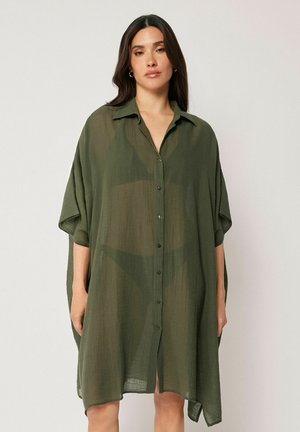 Kvinde med langt mørkt hår iført en løs, gennemsigtig olivengrøn kjole med knapper over sort undertøj, stående mod en lys baggrund.