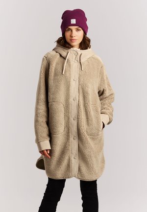 Jonge vrouw die een beige fleecejas draagt met grote zakken en een capuchon, gecombineerd met een paars mutsje en een zwarte broek, staand tegen een effen achtergrond.