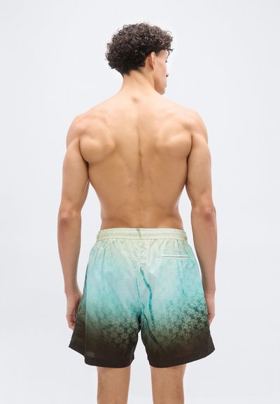 Άνδρας με σγουρά μαλλιά, χωρίς μπλούζα, γυρισμένος από πίσω, φοράει μαγιό με μοτίβο από πράσινο σε καφέ χρώμα, πάνω σε απλό φωτεινό φόντο.