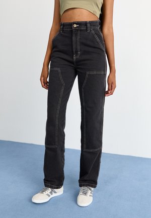 Pantalones de mezclilla negra con costuras blancas contrastantes, piernas rectas, bolsillos laterales y un cierre de botón en la cintura.