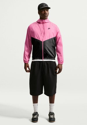 Mann trägt schwarze Kappe, pink-schwarze Nike Zip-Up-Jacke, schwarze lange Shorts, weiße Socken und schwarze Nike Sneakers und steht vor weißem Hintergrund.