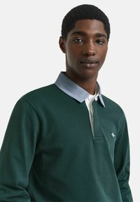 Junger schwarzer Mann, der ein dunkelgrünes Langarm-Polohemd mit einem hellblauen Kragen trägt und ruhig in die Kamera schaut, vor einem einfarbigen Hintergrund.