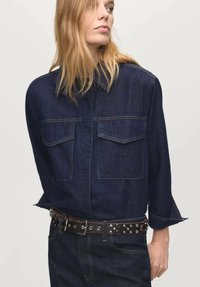 Camicia di jeans blu scuro con maniche lunghe, due tasche frontali e una patta con bottoni. Abbinata a una cintura nera con borchie argentate.